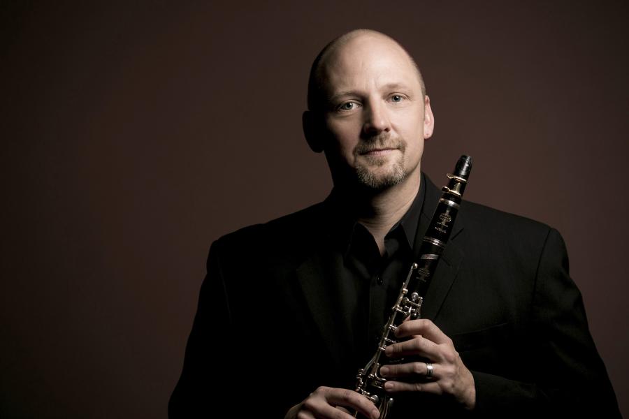 A man wearing a black suit holds a clarinet and smiles.