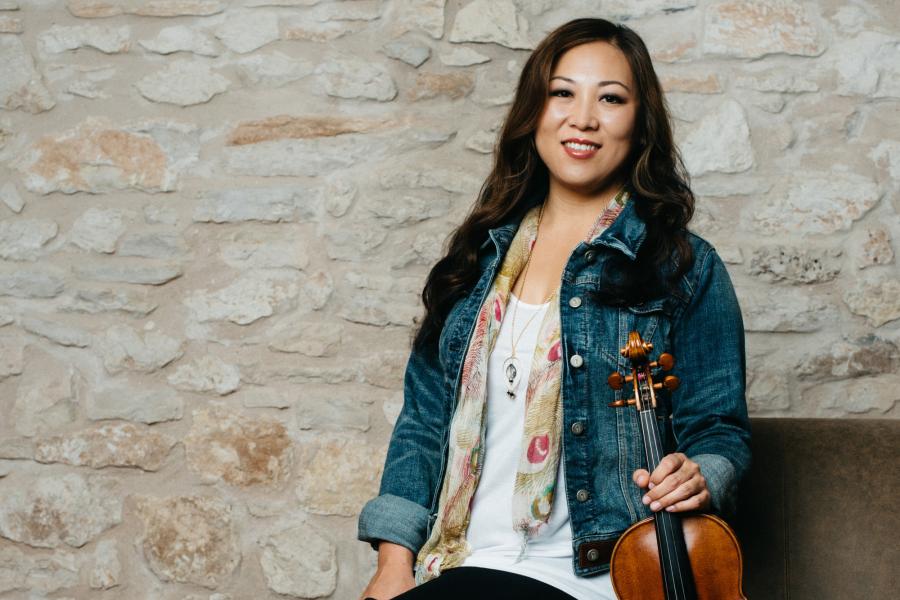 An Asian woman wearing a white shirt and jean jacket smiles and holds her violin.