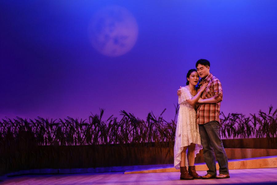A man and woman embrace under a full moon background on the opera stage