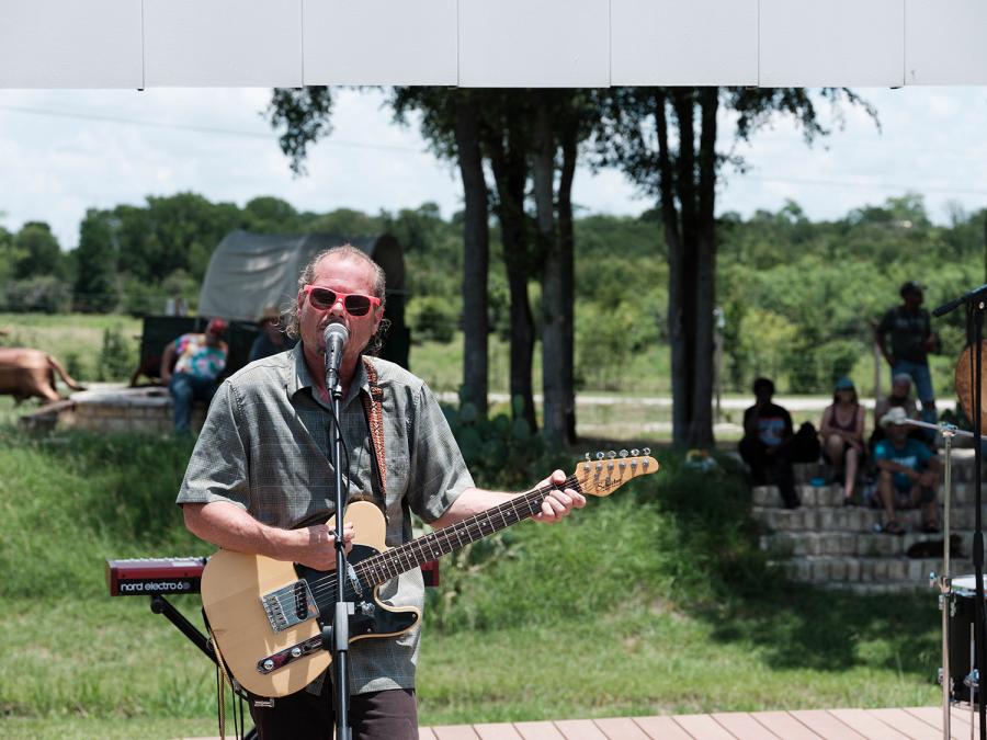 A Man in Pink sunglasses play Electric Guitar and Sings into a microphone on an outdoor stage