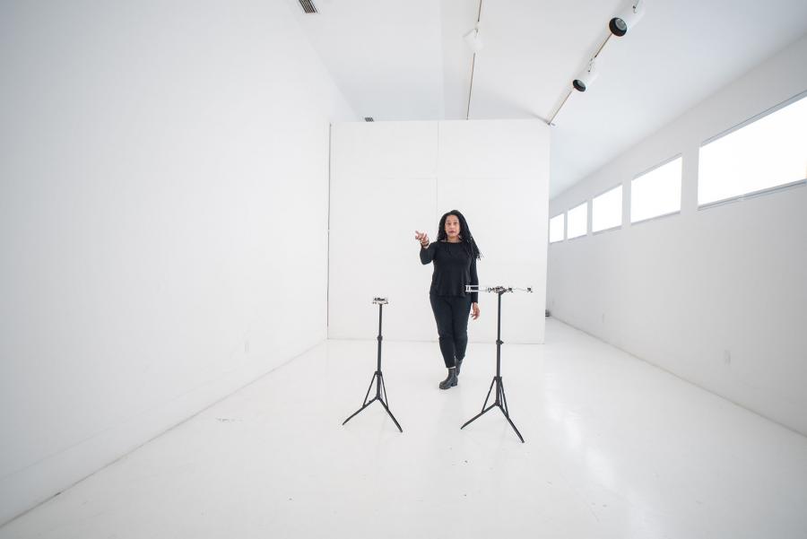Pamela Z stands confidently alone in a large all-white room with two microphones on stands in front of her.