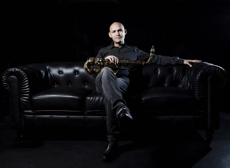 Miguel Zenón sits in a dark room on a black sofa with his saxophone across his lap