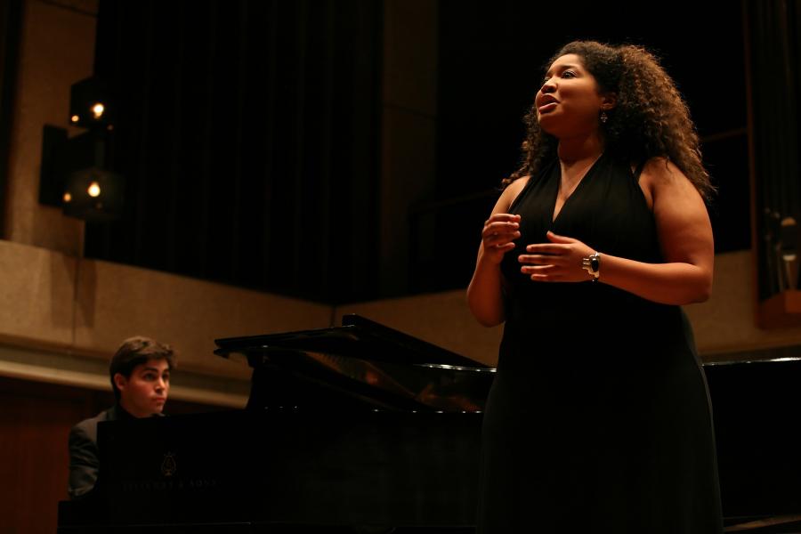 A Black woman sings while standing in front of a grand piano