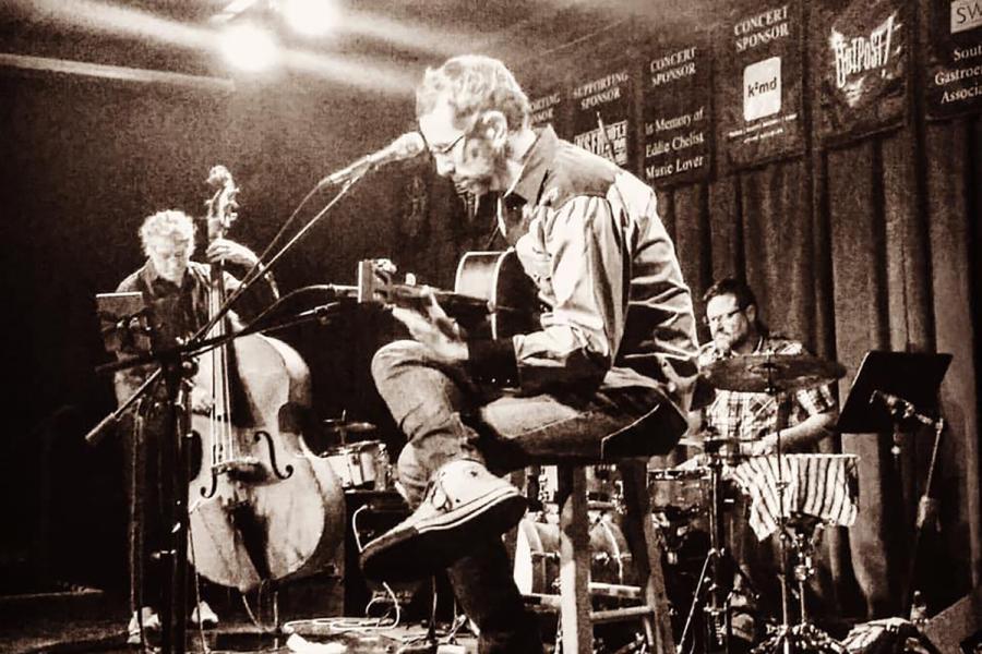 A sepia-tinted photo of a drummer, an upright bass player, and a guitarist performing on stage.