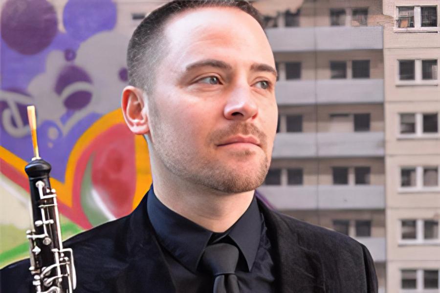 A man stands in front of a building with a mural. He is holding an oboe.