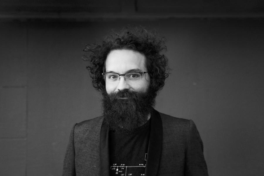 A black and white headshot of Thomas Echols with a long beard, glasses, and a t-shirt with a blazer.