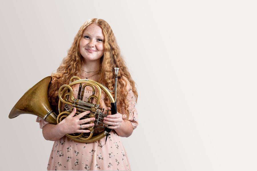 a horn player smiles warmly into the camera while holding her instrument