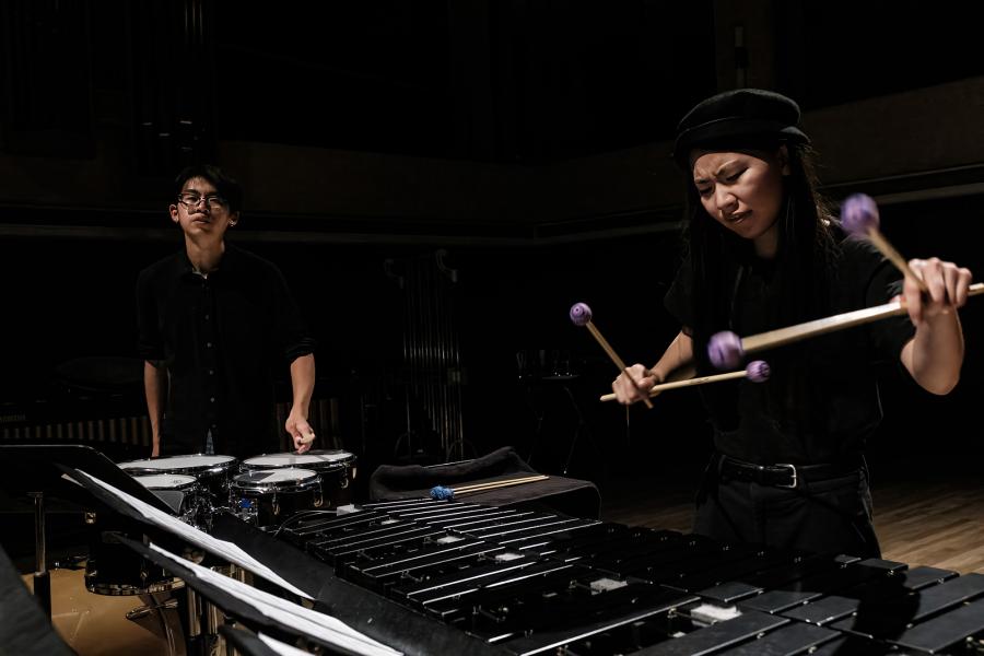 Two students play percussion instruments in a dimly lit hall.