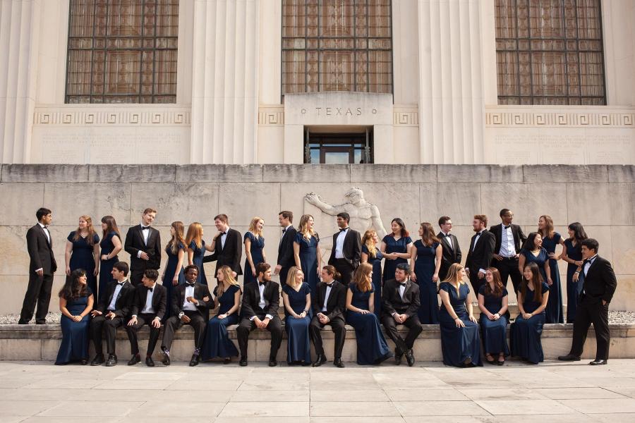 A large group of students dressed in formal attire posed casually in front of a University of Texas building. 