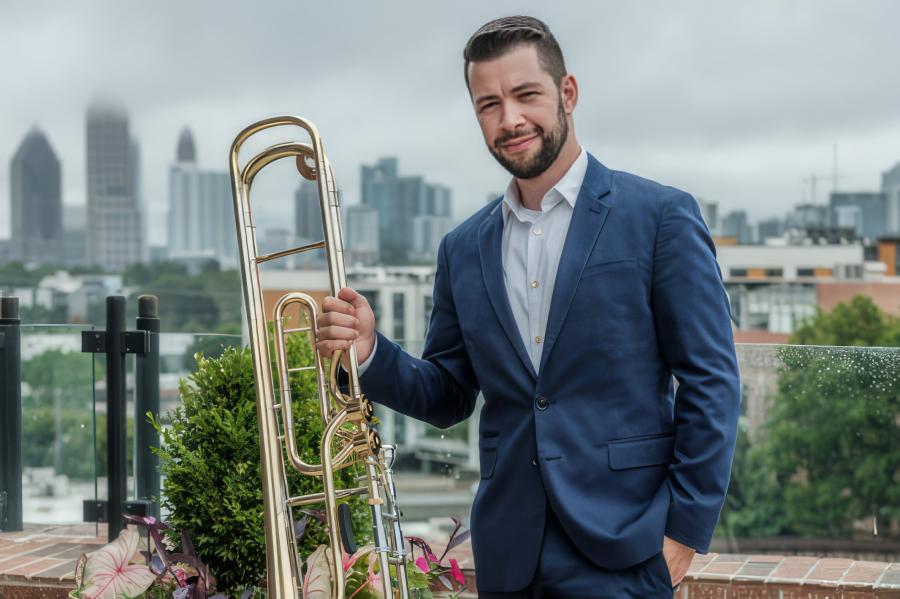 Brian Hecht, dressed in a blue suit, stands with his bass trombone, a city skyline is in the background