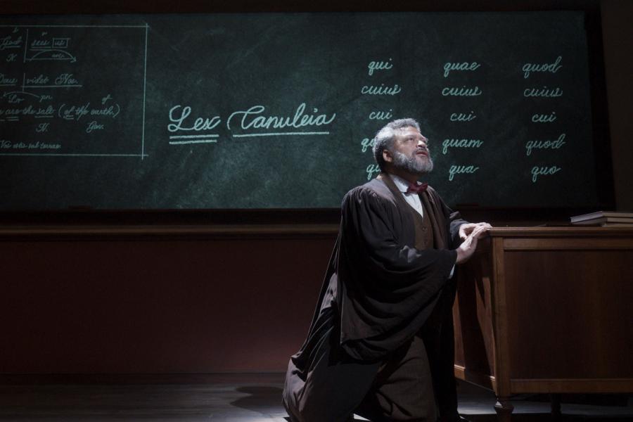 A still from the film Goodbye, Mr. Chips, showing a man kneeling beside a desk in front of a chalkboard.