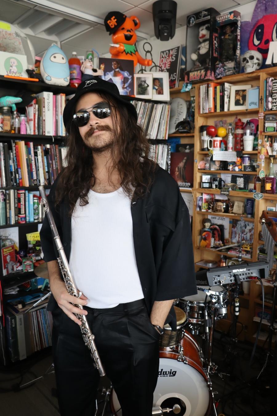 Paolo Santos holds his flute wearing sunglasses in front of the Tiny Desk set at NPR.