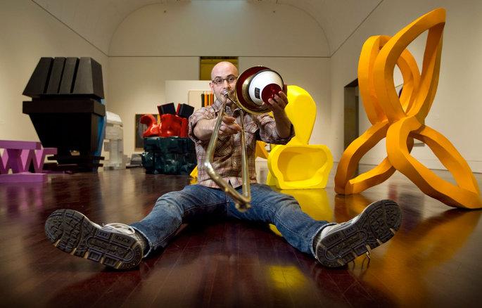 Steven Parker sits on the floor of a modern sculpture gallery, feet stretched in front of him, playing his trombone with a mute