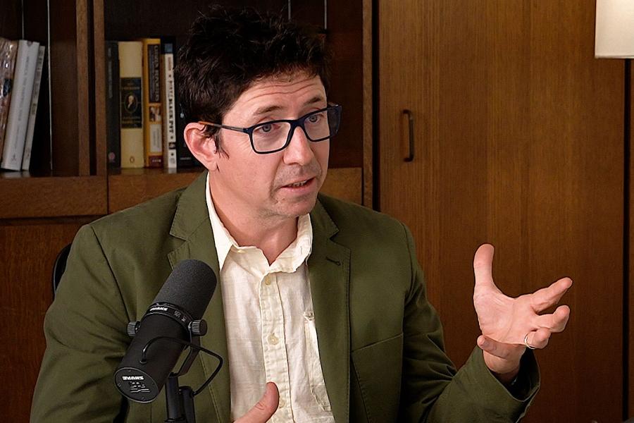 Sam Lipman sitting behind a mic with a bookshelf in the background