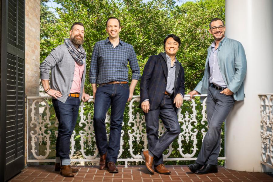 Four men wearing jeans smile and lean against decorative white railing on a porch. 