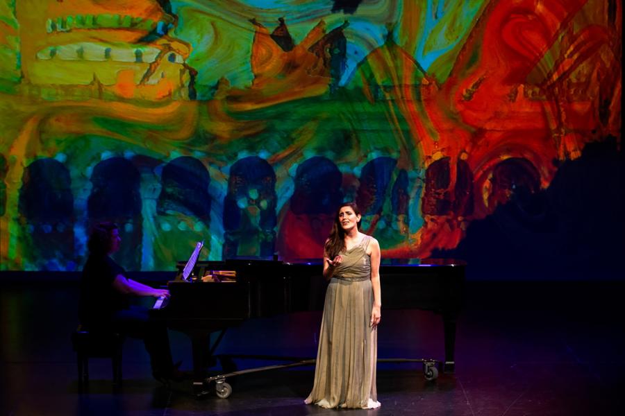 A singer performing on stage in front of a piano and a colorful abstract projected background