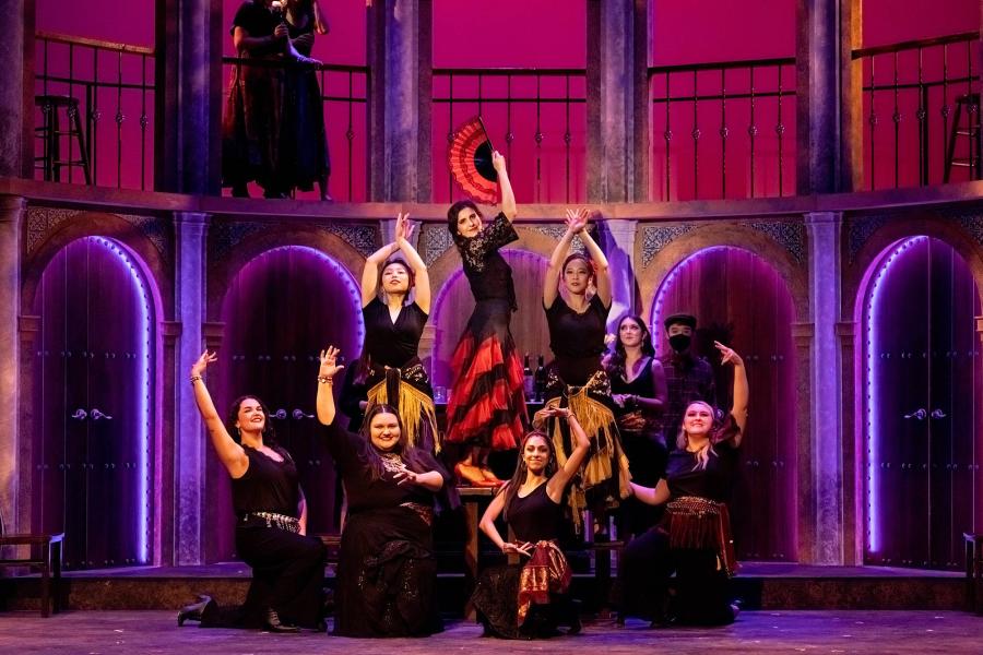 a group of dancers pose in an ornate 3-tier pyramid, with Carmen at the top, displaying a  hand fan.