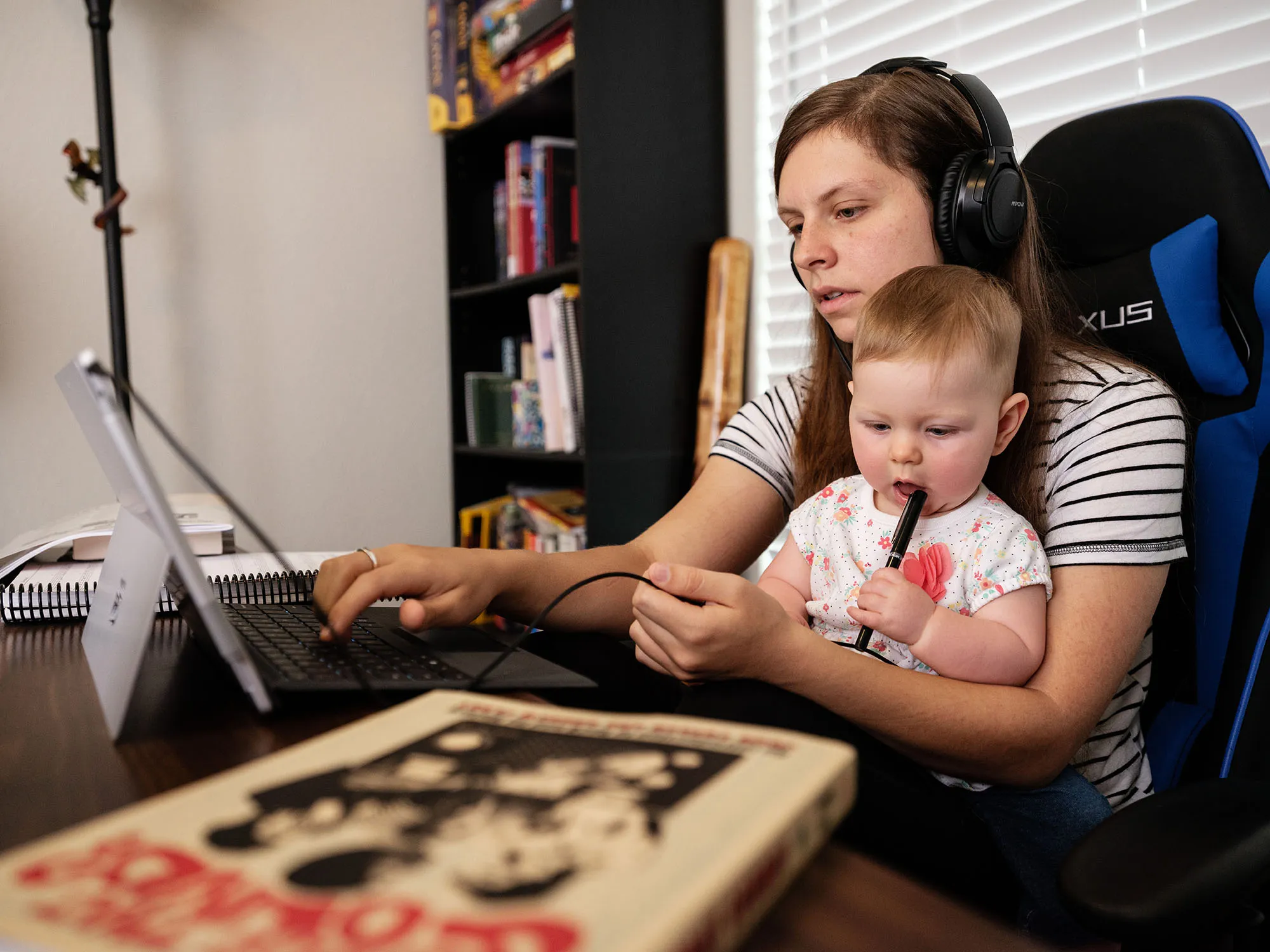 A student sits in a home office at her desk,, with a toddler on her lap.