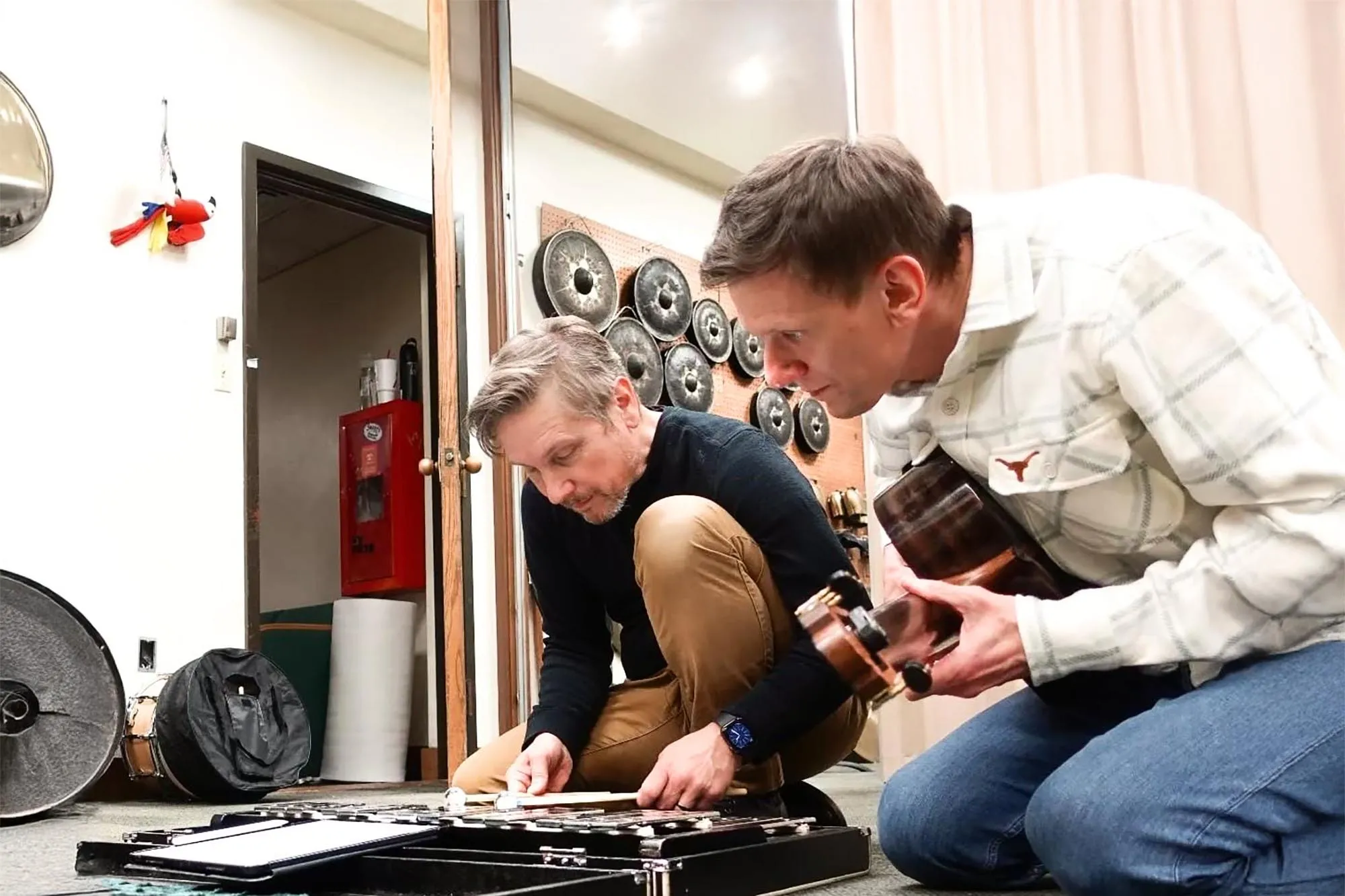 Burritt and Pasieczny kneel on the ground surrounded by percussion equipment inspecting a pedal board, while Pasieczny holds his guitar.