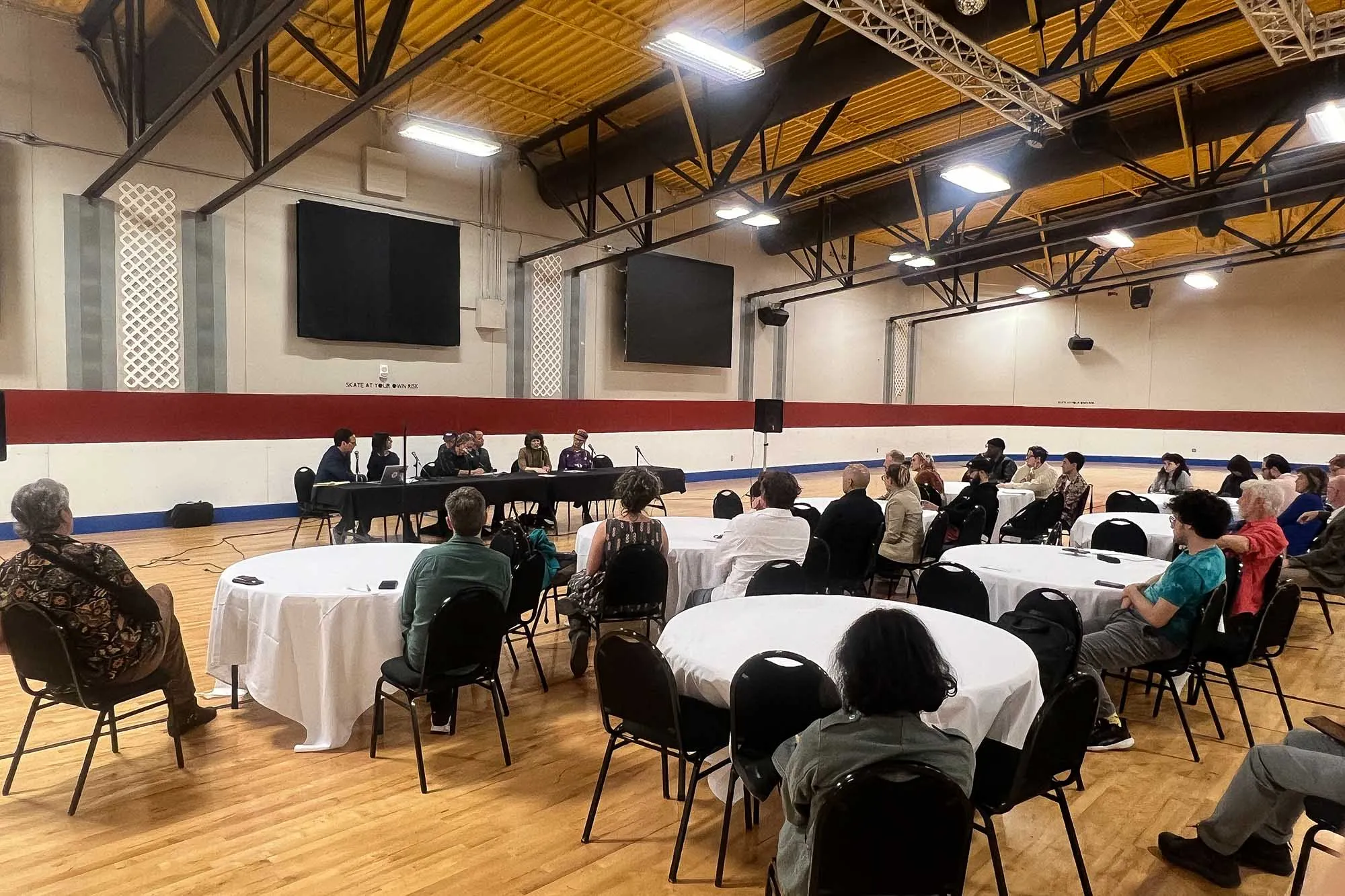 A large group of people sit at tables and listen to a presenter