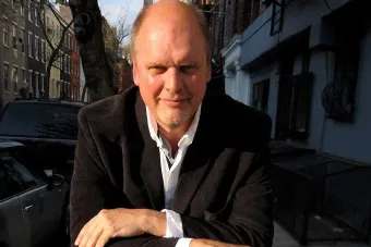 A white man in a white shirt and black jacket sits outside with his arms crossed and smiles.