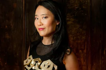 An Asian woman wearing a black and gold tank top and pink lipstick stands in front of a brown, scratched up door.