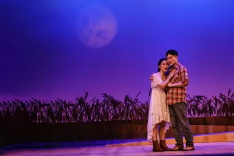 A man and woman embrace under a full moon background on the opera stage