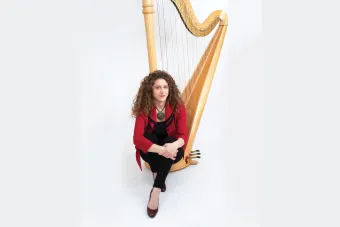 A woman in a red jacket sits at the foot of a harp.