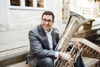 Benavidez sits on stairs outside a building with his tuba on his lap. 