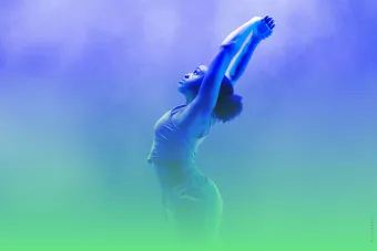 A dancer lifts her arms and arches her back in a hazy green and blue smoky background.