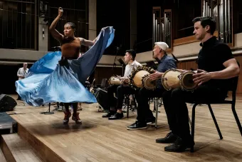 A woman in a blue skirt dancing on a stage in a well lit space in front of 3 seated men playing hand drums..