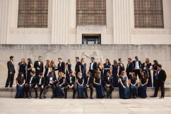 A large group of students dressed in formal attire posed casually in front of a University of Texas building.