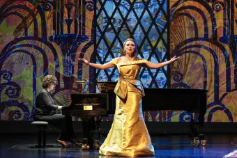 A singer in a formal yellow ball gown performs on stage with her arms outstretched, as a pianist accompanies behind her. 