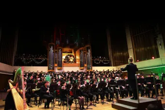The University of Texas Wind Ensemble plays with combined choirs, conducted by J.D. Burnett. Bates Recital Hall is decorated with lights and Christmas garland.