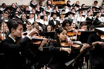The first violin section of the University of Texas Symphony Orchestra plays, foregrounded by University of Texas choral singers.