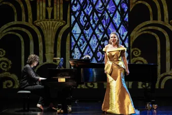 Dr. Tamar Sanikidze plays piano while soprano Kara Covey sings wearing a gold gown. A stained glass window and pillars are projected behind them.