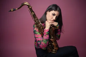 Melissa Aldana sits for the camera, holding her tenor saxophone. 