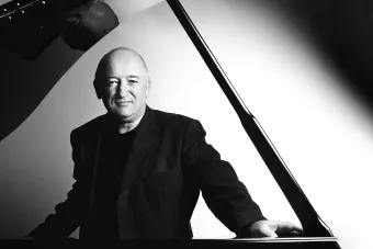 John O'Conor posed in the bend of a concert grand piano, looking at the viewer through the piano