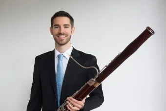 Drew Pattison holds his bassoon wearing a black suit with a blue tie.
