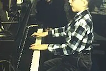 A very young Gregory Allen plays an upright piano while a woman looks on. 