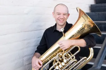 Tuba player Scott Sutherland sits on a staircase holding his instrument in his lap.