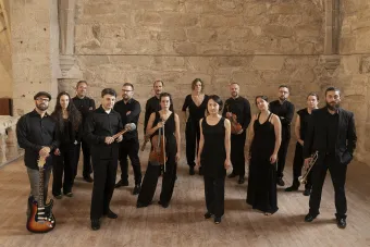 Members of Vertixe Sonora Ensemble stand in a stone room, dressed in black, holding their instruments.