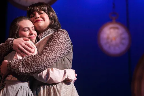 Two women embrace each other with heads close and eyes closed. In the background out of focus, are several large clocks hanging by chains.