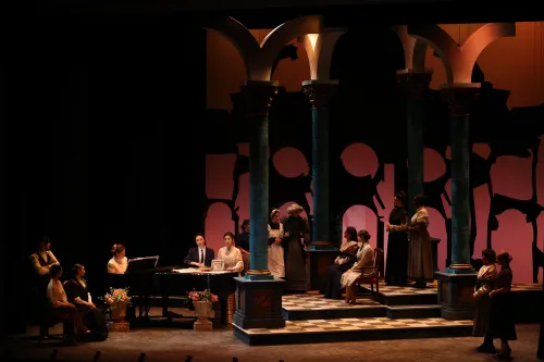 performs stand around a stage that is dimly lit in a soft pink glow on a large set with many tables and pillars.  