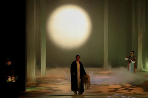 A shadowy figure stands at the center of a fog-filled stage, with a soft glowy moon in the background.