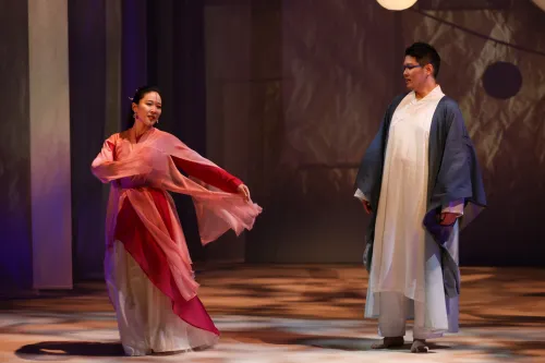 a woman dress in a flowing pink robe dances on stage while a man looks on.