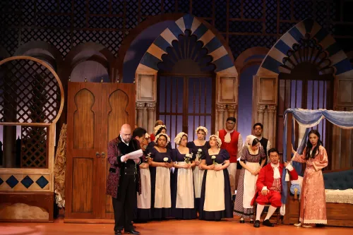 a large cast assmebles on bedroom set, as one man reads from a scroll, center stage. 