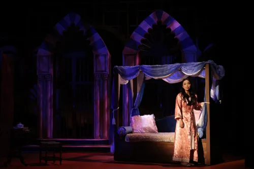 A woman in a nightgown stands holding a bedpost in a dimly lit bedroom set, she is singularly lit with a spotlight. 