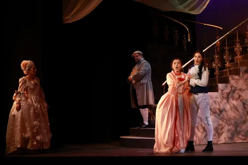 A man and woman in Regency attire embrace one another at the bottom of a grand staircase, while other cast members look on, somberly. 