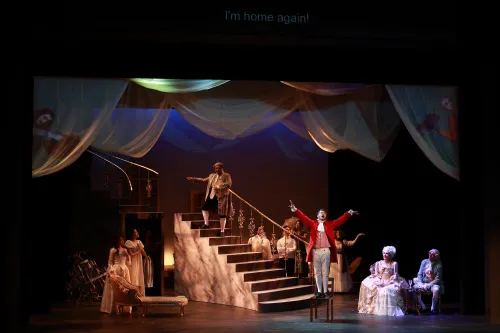 The cast of Ghost of Versailles are stage on and around a grand staircase at center stage, a man in a bright red overcoat is performing with his arms outstretched. 